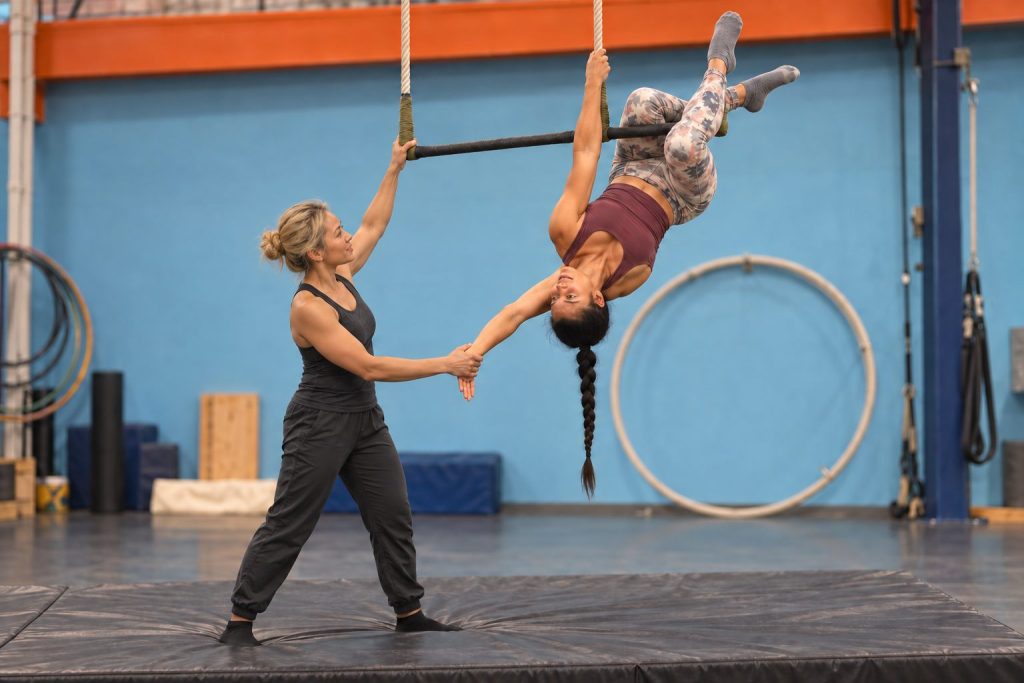 A student hangs upside down from a static trapeze by her knees while a coach steadies her hand